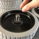 Floor scrubber gray roller wheel, anti-slip tread surface, hand installing metal shaft, stacked wheels in background.